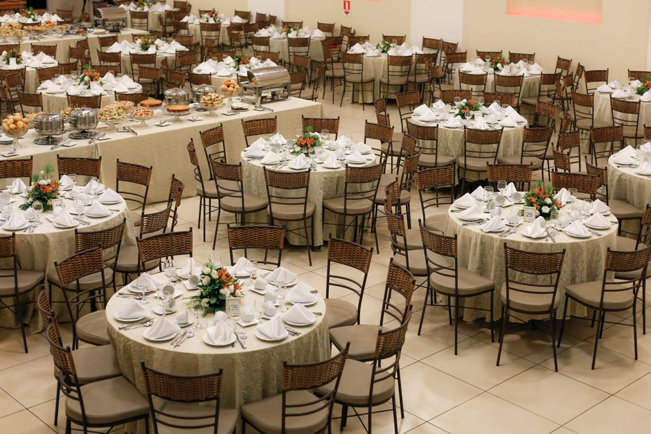 A spacious banquet hall set up for a formal event features multiple round tables draped with cream-colored tablecloths. Each table is elegantly arranged with white napkins folded into a standing fan s