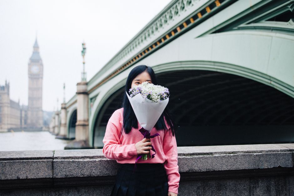 A young woman with long black hair, wearing a pink sweater and black skirt, holds a bouquet of fresh flowers wrapped in white paper and tied with a dark purple ribbon. The bouquet includes small white