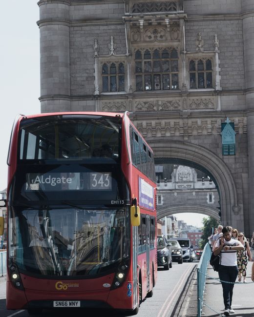 A red double-decker bus displaying the route to Aldgate, with the destination sign visible at the front, passing underneath the historic Tower of London’s stone gateway with ornate architectural det