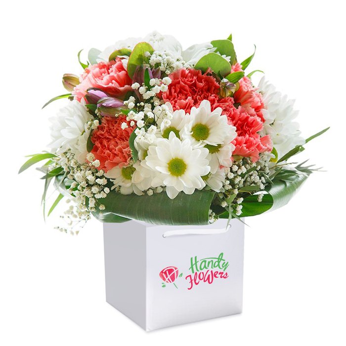 This expertly constructed bouquet, rising with elegance from a square white gift bag, reflects Aldgate's unique blend of contemporary life and historic charm. The heart of the arrangement is a profusion of peach, coral, and vivid red carnations-each ruffled petal unfurling with soft grandeur and a tactile richness that delights the senses. Interspersed within the bouquet, radiant white daisies with vivid green-gold centres inject vibrance, drawing parallels to the green spaces scattered throughout the Aldgate area. Wisps of white baby's breath float gently among the main blooms, their fine-detail filaments imparting a generous breath of freshness and airiness. On the bouquet's left flank, a handful of closed violet buds softly introduce intrigue and invite a closer look. Swirling around the composition, glossy, deep green leaves form a protective and elegant base, echoing the balance of nature with city life found near Aldgate Square or along Leman Street. The white gift bag, sturdy and purposeful with crisp white handles, makes the bouquet easily portable-suited to local gifting and thoughtful deliveries. Captured against a flawless white background, every detail and texture is illuminated, highlighting the bouquet's freshness and expert arrangement. With every stem thoughtfully placed by skilled local hands, this bouquet is ideal for meaningful occasions-be it birthdays, anniversaries, or brightening a loved one's workspace near the City.